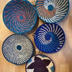 May include: Five round woven baskets in shades of blue, teal, and purple, with spiral and star patterns. The baskets are displayed on a light wood surface. Each basket has a unique design, with accents in orange, white, and turquoise.