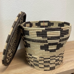 May include: A woven basket with a lid, featuring a geometric pattern in shades of brown and beige. The basket is round with a slightly flared shape, and the lid is partially open, revealing the interior. The basket is sitting on a wooden surface.