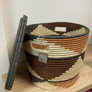 May include: A woven basket with a geometric pattern in shades of brown, beige, and black. The basket has a handle and a tag attached. The handle is a dark gray color. The basket is sitting on a wooden surface.