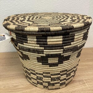 May include: A woven basket with a lid, featuring a geometric pattern in shades of brown and beige. The basket has a cylindrical shape and is made of natural fibers. The lid has a concentric circle design.