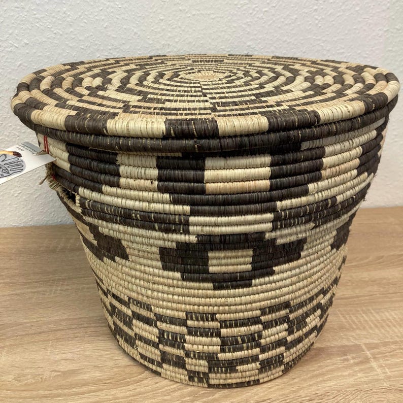 May include: A woven basket with a lid, featuring a geometric pattern in shades of brown and beige. The basket has a cylindrical shape and is made of natural fibers. The lid has a concentric circle design.