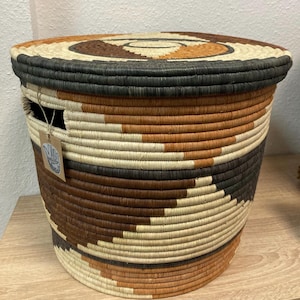 May include: Woven basket with a lid, featuring a spiral design on top. The basket is cylindrical, with alternating bands of brown, black, and natural fibers. A small tag hangs from the side. This decorative storage basket is suitable for various uses.