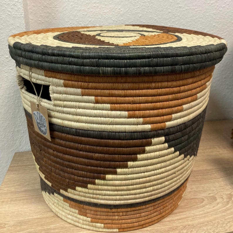 May include: Woven basket with a lid, featuring a spiral design on top. The basket is cylindrical, with alternating bands of brown, black, and natural fibers. A small tag hangs from the side. This decorative storage basket is suitable for various uses.