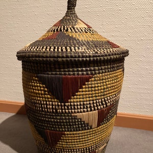 May include: A woven basket with a lid, featuring a conical top and a cylindrical body. The basket is adorned with a geometric pattern in shades of brown, tan, and yellow. The lid has a small, woven handle.