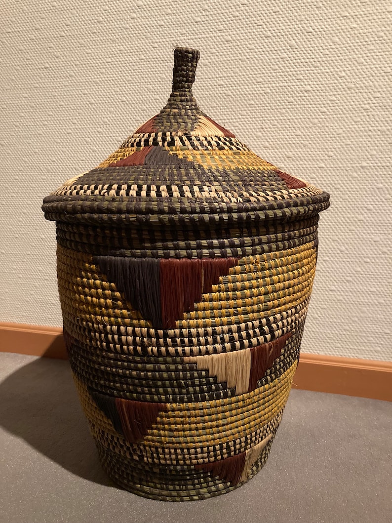 May include: A woven basket with a lid, featuring a conical top and a cylindrical body. The basket is adorned with a geometric pattern in shades of brown, tan, and yellow. The lid has a small, woven handle.