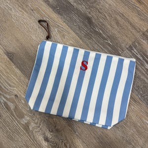 May include: A rectangular canvas pouch with blue and white vertical stripes. The pouch has a red letter "S" on the front and a brown leather loop attached to the zipper. The pouch is lying on a wooden surface.