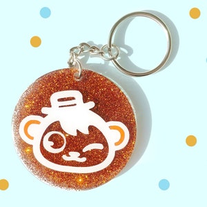 May include: A round, copper-colored glitter keychain with a white cartoon animal wearing a hat. The animal has large eyes and a winking expression.