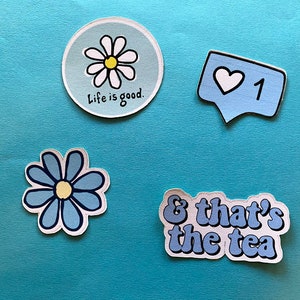 Puede incluir: Cuatro pegatinas sobre un fondo azul. Una es una margarita con el texto "Life is good". Otra es una burbuja de di&aacute;logo con un coraz&oacute;n y el n&uacute;mero "1". Hay una pegatina de flor azul y una pegatina que dice "& that's the tea."