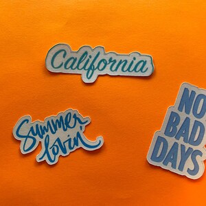 Puede incluir: Tres pegatinas con bordes blancos y texto azul sobre un fondo naranja. Las pegatinas dicen "California", "Summer lovin" y "No bad days". Las pegatinas son probablemente para decorar art&iacute;culos.