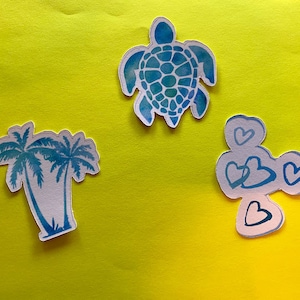 Puede incluir: Tres pegatinas troqueladas sobre un fondo amarillo. Las pegatinas incluyen una tortuga azul de acuarela, un grupo de tres corazones azules y un par de palmeras. Las pegatinas tienen un borde blanco.