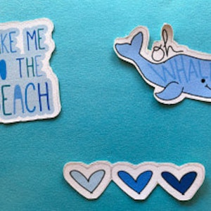 Puede incluir: Tres pegatinas sobre un fondo azul. La primera dice "TAKE ME TO THE BEACH" en texto azul. La segunda es una ballena azul con la frase "oh whale" escrita encima. La tercera tiene tres corazones azules.