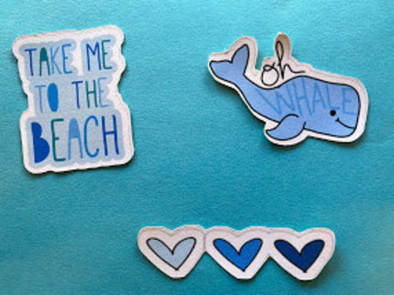 Puede incluir: Tres pegatinas sobre un fondo azul. La primera dice "TAKE ME TO THE BEACH" en texto azul. La segunda es una ballena azul con la frase "oh whale" escrita encima. La tercera tiene tres corazones azules.