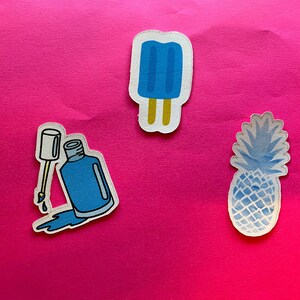Puede incluir: Tres pegatinas sobre un fondo rosa brillante. Las pegatinas incluyen un helado azul en un palo, un frasco de esmalte de u&ntilde;as azul con un pincel y una pi&ntilde;a azul. El esmalte de u&ntilde;as se est&aacute; derramando de la botella.