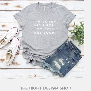 May include: Gray t-shirt with the text "I'M SORRY DID I ROLL MY EYES OUT LOUD?" in white. The shirt is laid flat on a white wooden surface with a pair of white sneakers and denim shorts.