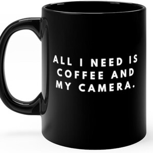 May include: Black coffee mug with white text that reads "All I need is coffee and my camera."