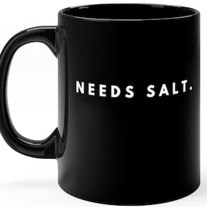 May include: Black ceramic coffee mug with a curved handle. The mug features the white text "NEEDS SALT." on the side. The mug is designed for hot or cold beverages.