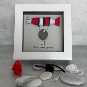 May include: A framed artwork featuring a small pebble person holding up a red and white striped yarn football. The text "Manchester United" is written below the pebble person.