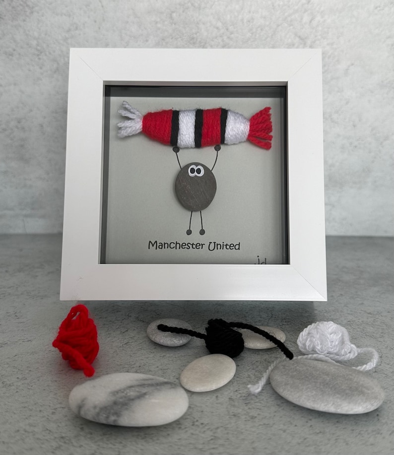 May include: A framed artwork featuring a small pebble person holding up a red and white striped yarn football. The text "Manchester United" is written below the pebble person.