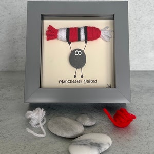 May include: A framed artwork featuring a gray pebble with googly eyes holding up a red and white yarn football. The text "Manchester United" is written below the pebble.