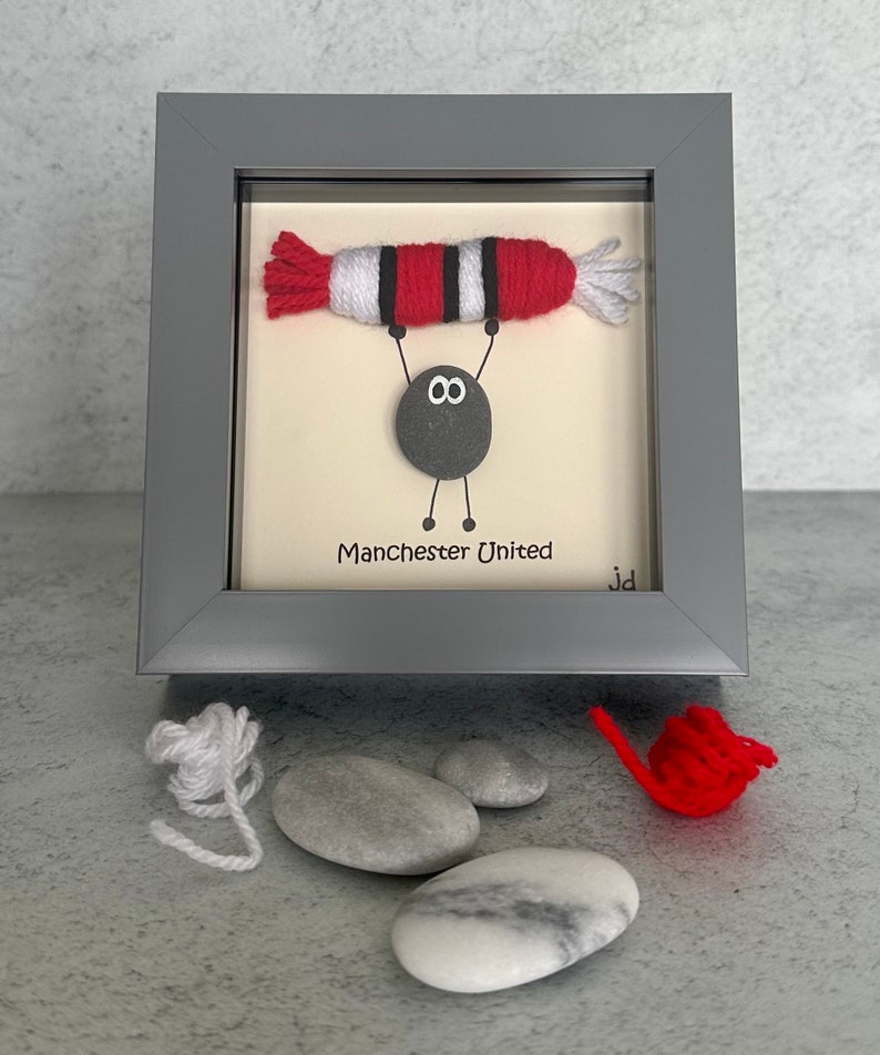 May include: A framed artwork featuring a gray pebble with googly eyes holding up a red and white yarn football. The text "Manchester United" is written below the pebble.