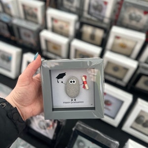 May include: A grey framed picture featuring a painted rock wearing a graduation cap and holding a diploma. The rock has googly eyes and a smile. The text "Happy Graduation!" is written below the rock.