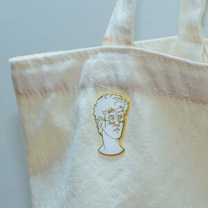 May include: A white enamel pin with a gold outline depicting a profile of a person's face. The pin is attached to a white canvas tote bag.