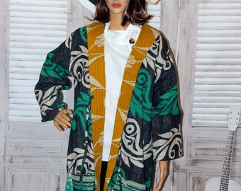 May include: A black and green kantha quilted jacket with a white and gold floral pattern. The jacket is open and has a white shirt underneath. The model is wearing a black hat.