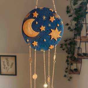 Könnte beinhalten: Hängedekoration mit Himmelsmotiv, bestehend aus einer blauen Scheibe mit goldenen Akzenten. Das Design umfasst einen Mond, eine Sonne und Sterne. Goldene Ketten mit dekorativen Elementen und Kristallanhängern hängen.