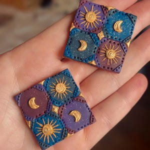 Peut inclure: Deux boucles d'oreilles carrées faites à la main avec des motifs célestes. Chaque boucle d'oreille présente un soleil et un croissant de lune dorés sur un fond bleu, violet et marron. Les bords sont soulignés en or, créant un accessoire artistique unique.