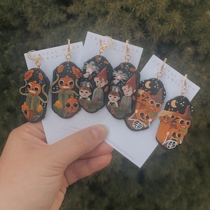 Puede incluir: Pendientes de cerámica hechos a mano con diseños caprichosos. Los pendientes muestran personajes y escenas en tonos naranja, marrón y negro, con ganchos dorados. El texto "CRYSTAL MOON CERAMICS" es visible en las tarjetas de respaldo.