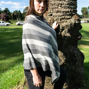 Op de afbeelding: Een grijs en wit gestreepte poncho. De poncho wordt over de schouders gedragen. De foto is buiten genomen op een zonnige dag met een blauwe lucht en groen gras op de achtergrond. De poncho is gemaakt van een zacht materiaal.