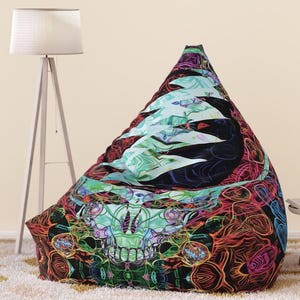 May include: A large, triangular beanbag chair with a colorful, psychedelic skull design. The beanbag features a mix of red, green, blue, and black patterns. The chair is on a cream-colored rug.