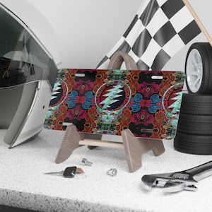 May include: A license plate featuring a colorful, psychedelic design with a skull and lightning bolt motif. The plate is displayed on a wooden stand, with a racing flag and tires in the background. A helmet, wrench, and keys are also visible.