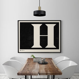May include: A black and white poster with a large white letter H on a black background. The poster is framed in black and hanging on a white wall above a wooden table with white chairs.