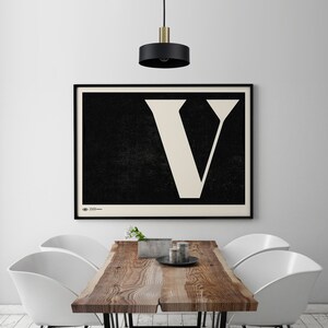May include: A black and white poster featuring a large white letter V against a black background. The poster is framed in black and hangs on a white wall above a wooden table with white chairs.