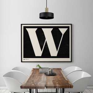 Puede incluir: Un póster en blanco y negro con una gran letra W blanca sobre un fondo negro. El póster está enmarcado en negro y colgado en una pared blanca sobre una mesa de madera con sillas blancas.