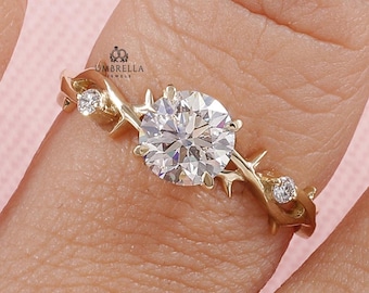 Könnte beinhalten: Ein Goldring mit einem großen runden Brillantschliff-Mittelstein, flankiert von zwei kleineren runden Steinen. Die Ringschiene ist mit einem strukturierten, dornigen Rankenmotiv gestaltet. Der kleine Text Umbrella Jewels ist sichtbar.