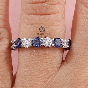 Könnte beinhalten: Ein silberner Ring mit abwechselnd runden, brillant geschliffenen weißen und blauen Edelsteinen. Der Ring ist mit mehreren Steinen besetzt, wodurch ein durchgehendes Band aus Glanz entsteht. Der Ring wird an einem Finger gezeigt.