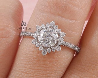 May include: A silver-toned ring featuring a large round center stone surrounded by a halo of smaller round stones in a starburst pattern. The band is thin and encrusted with tiny stones. The text UMBRELLA JEWELS is visible near the setting.