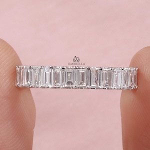 May include: A silver ring with a continuous row of rectangular, clear gemstones. The ring is held between two fingers against a soft pink background. The ring's design is simple and elegant, showcasing the sparkle of the gemstones.