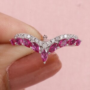 May include: A V-shaped ring featuring a row of marquise-cut pink stones alternating with a row of small round clear stones. The metal band is silver-toned. The text UMBRELLA JEWELS is printed in small black letters near the center of the ring.