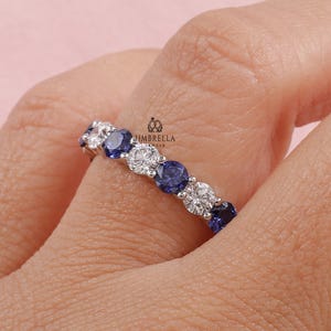 以下が含まれることがあります： 青と透明な宝石が交互に並んだシルバーリング。リングにはラウンドカットの石がセットされており、連続した色の帯を形成しています。リングは指にあり、ピンク色の背景です。