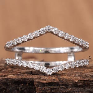 May include: A silver-toned ring guard featuring two chevron-shaped bands connected at the sides. Both bands are encrusted with small, clear, round-cut stones along the entire outer surface, creating a continuous sparkling effect.