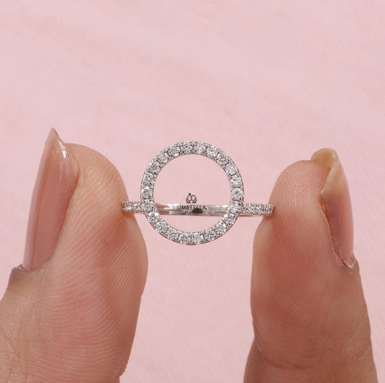 May include: A silver ring with a circular design, encrusted with small, sparkling stones. The ring features a delicate band and a central open circle, creating a modern and elegant look. The word "UMBRELLA" is engraved on the ring.