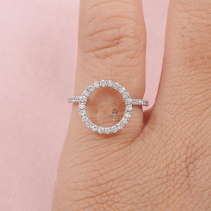 May include: A silver ring featuring a circular design encrusted with small, sparkling stones. The ring is set against a light pink background, showcasing the intricate details of the jewelry. The ring is a delicate piece of jewelry.