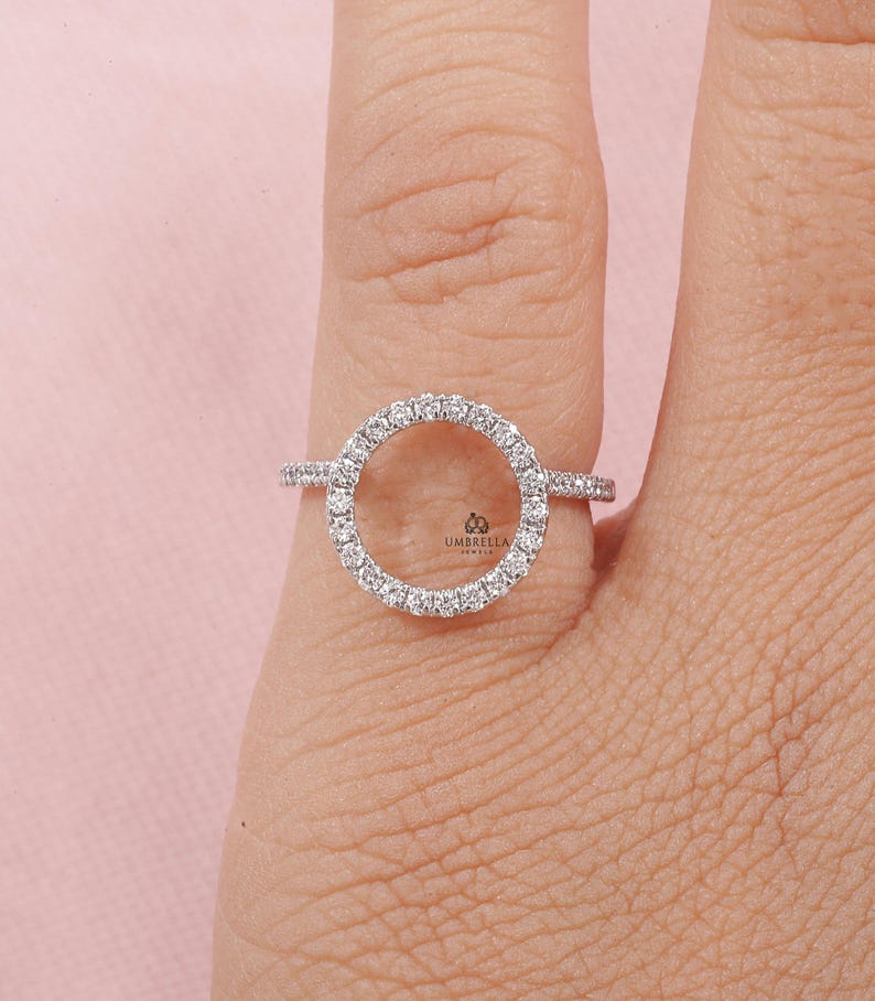 May include: A silver ring featuring a circular design encrusted with small, sparkling stones. The ring is set against a light pink background, showcasing the intricate details of the jewelry. The ring is a delicate piece of jewelry.