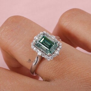 May include: A silver-toned ring featuring a large, emerald-cut green gemstone at the center, surrounded by a halo of small, round, clear stones. The band is smooth and metallic. The text UMBRELLA JEWELS is visible on the ring setting.