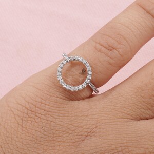 May include: A silver ring with a circular design, encrusted with small, clear gemstones. The ring is set on a finger against a soft pink background. The ring's design features a halo of gemstones around an open center.