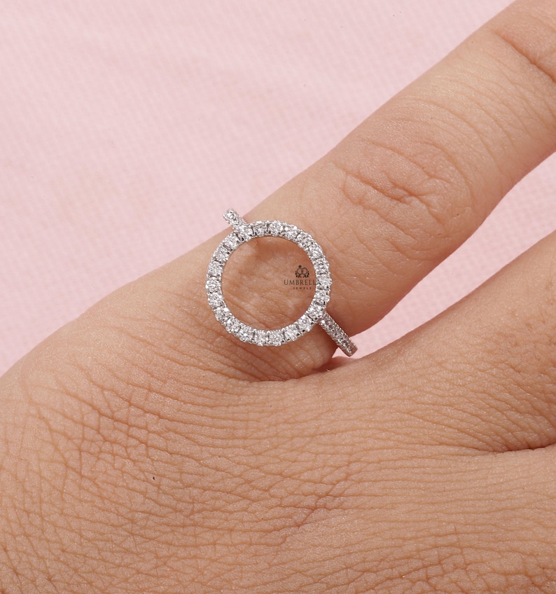May include: A silver ring with a circular design, encrusted with small, clear gemstones. The ring is set on a finger against a soft pink background. The ring's design features a halo of gemstones around an open center.