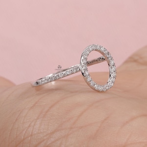 May include: A silver ring with a circular design, encrusted with small, sparkling stones. The band and the circle are both adorned with the same stones, creating a continuous, elegant look. The ring is set against a soft pink background.